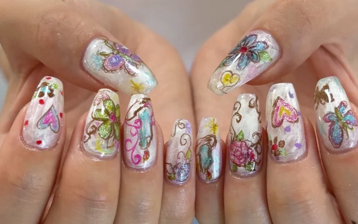 Hand-painted Ang:tt nail art with colorful flowers, hearts, and butterflies on pearly white nails.