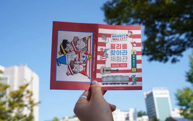 Hand holds Where’s Wally? Hongdae experience guide map under a blue sky.