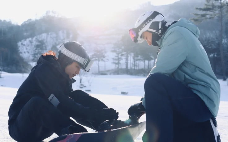 Instructor helping a snowboarder fasten bindings on a sunlit slope in Oak Valley.