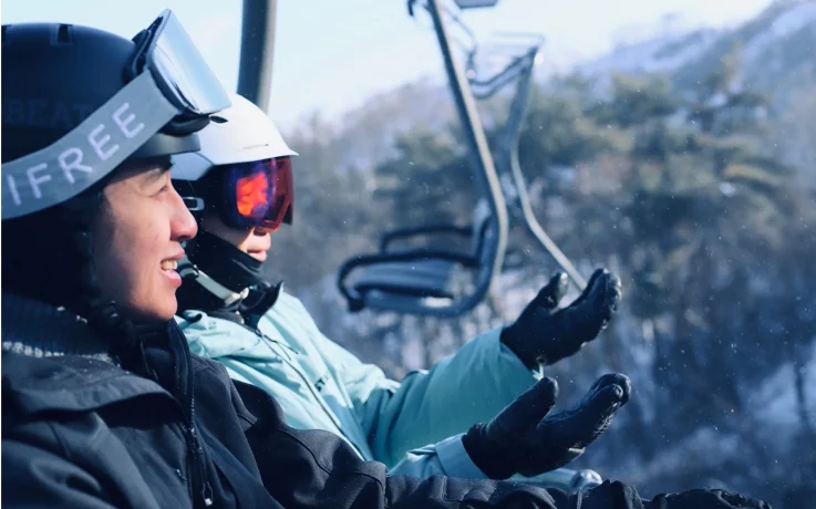 Two skiers chatting on a chairlift above a snow-dusted forest in Oak Valley.