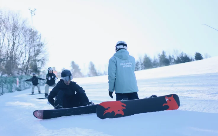 Oak Valley Ski & Board School instructor guiding a beginner snowboarder on a sunny gentle slope.