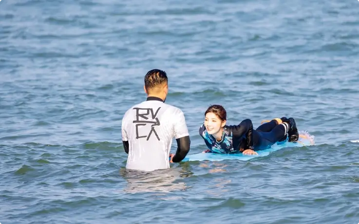 A surf instructor guides a student lying on a surfboard in the water.