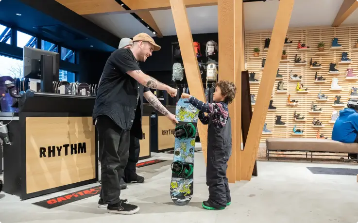 Friendly store clerk hands a snowboard to a child at a ski rental store.